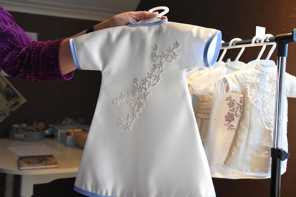 Hand som håller upp en liten vacker, vit klänning i babystorlek. Länk till webbnyhet.