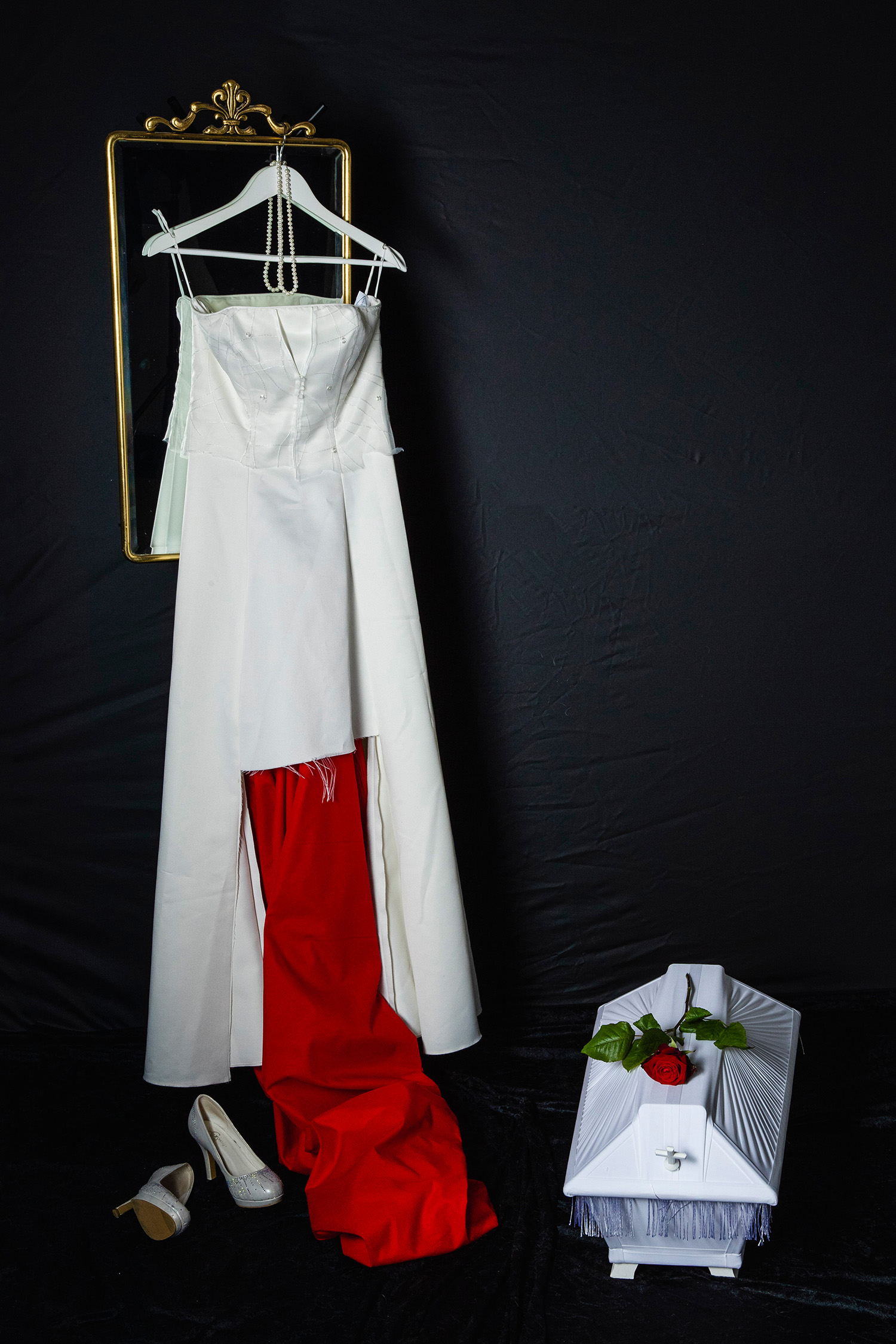 En sönderklippt brudklänning upphängd på en spegel med guldram. På golvet finns en liten kista och ett par vita skor.