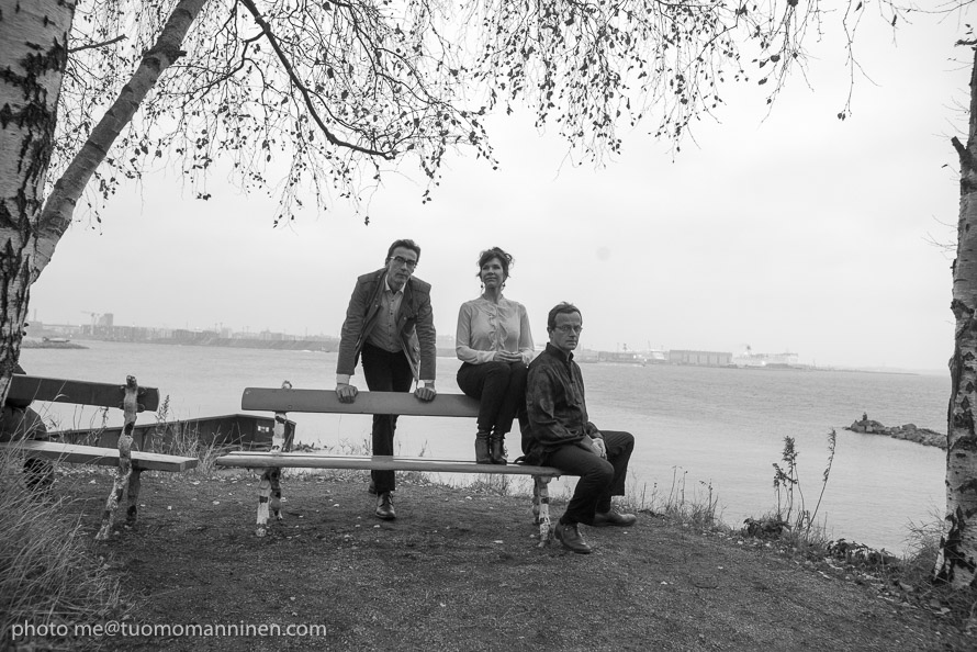Tre personer sitter och står kring en bänk i en park vid havsstranden.