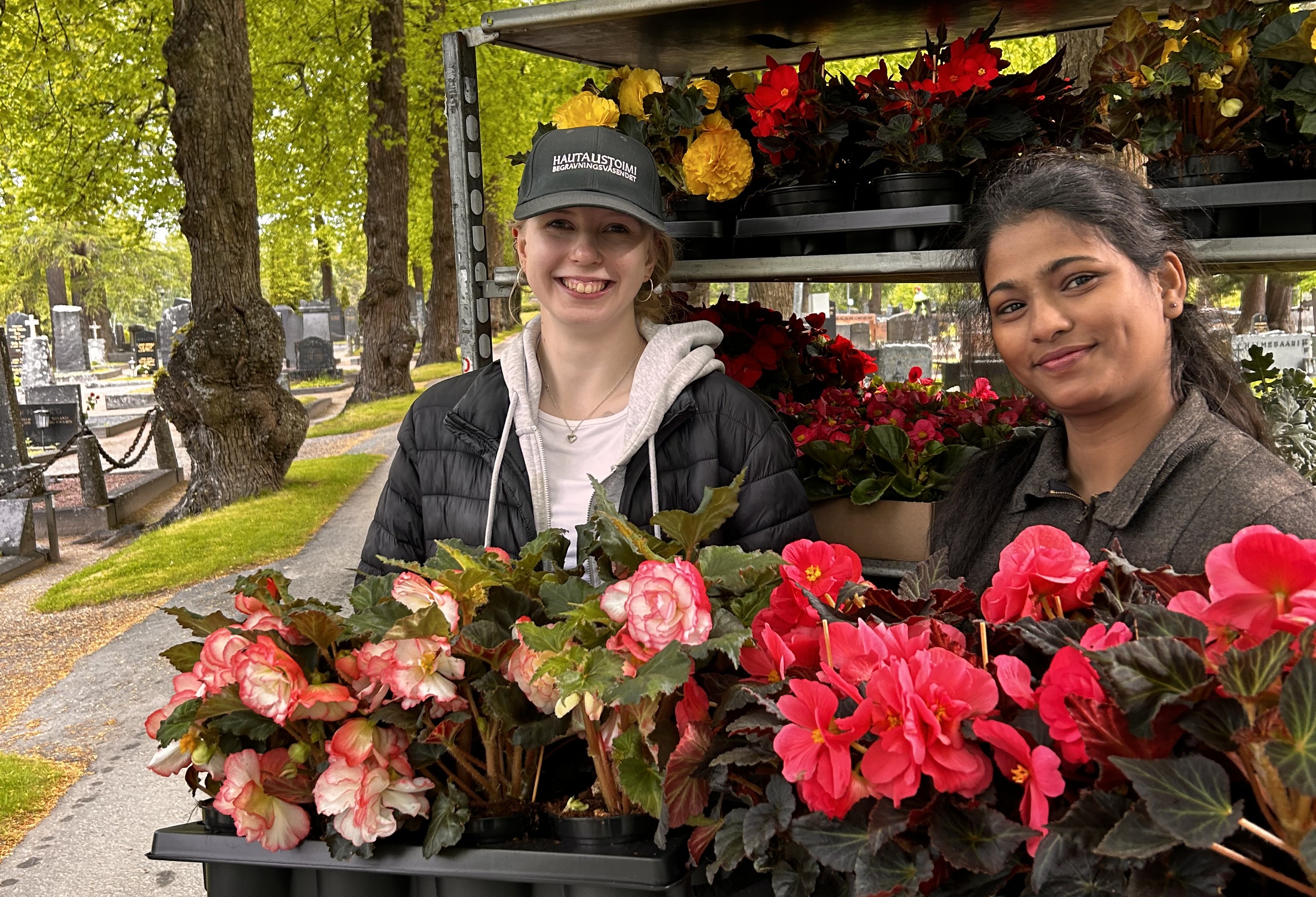 Två unga kvinnor på en begravningsplats håller lådor med blommor i sina famnar.