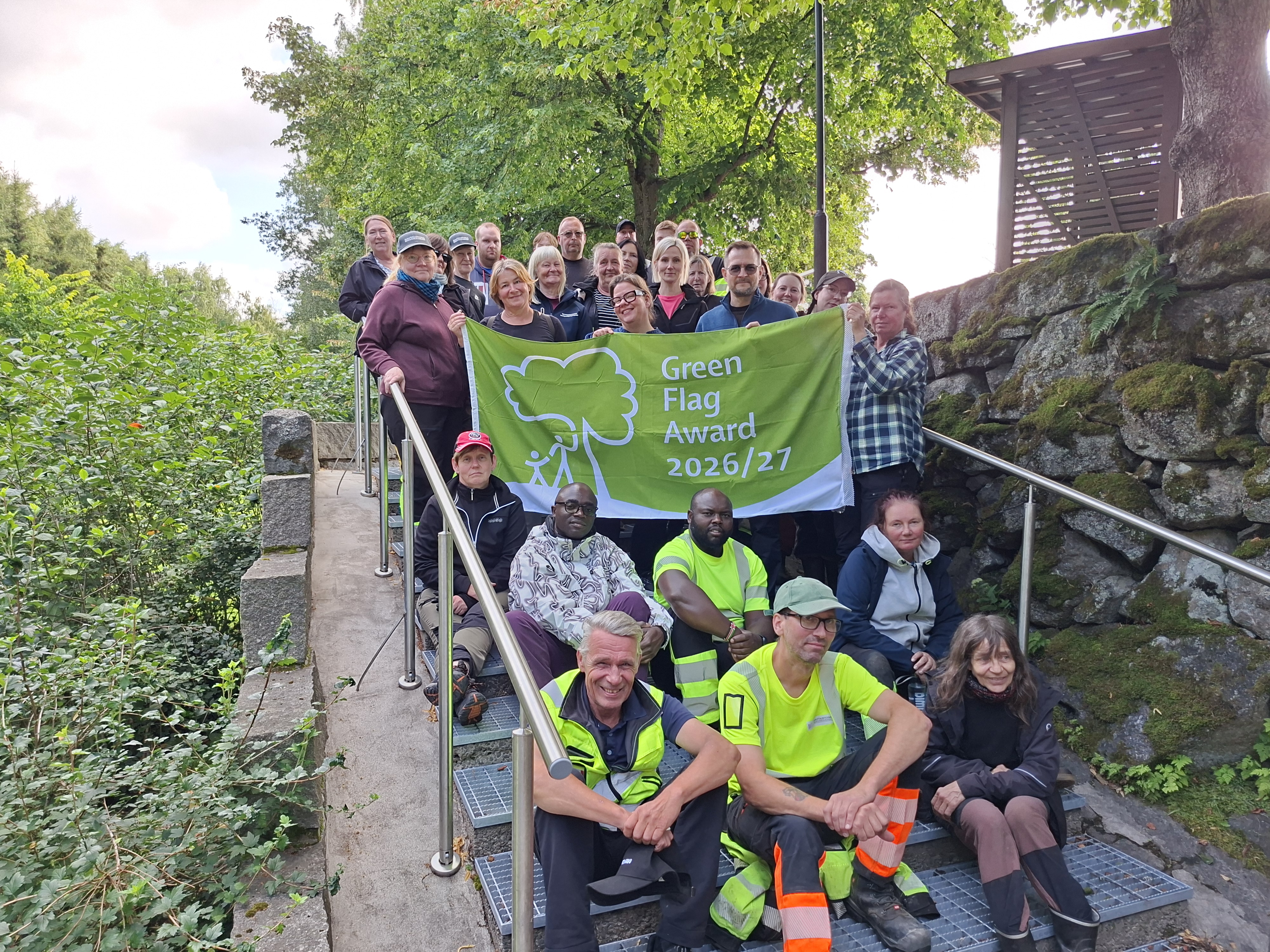 Grupp med människor står och sitter på en trappa utomhus. Tre personer håller i en flagga med texten: Green...