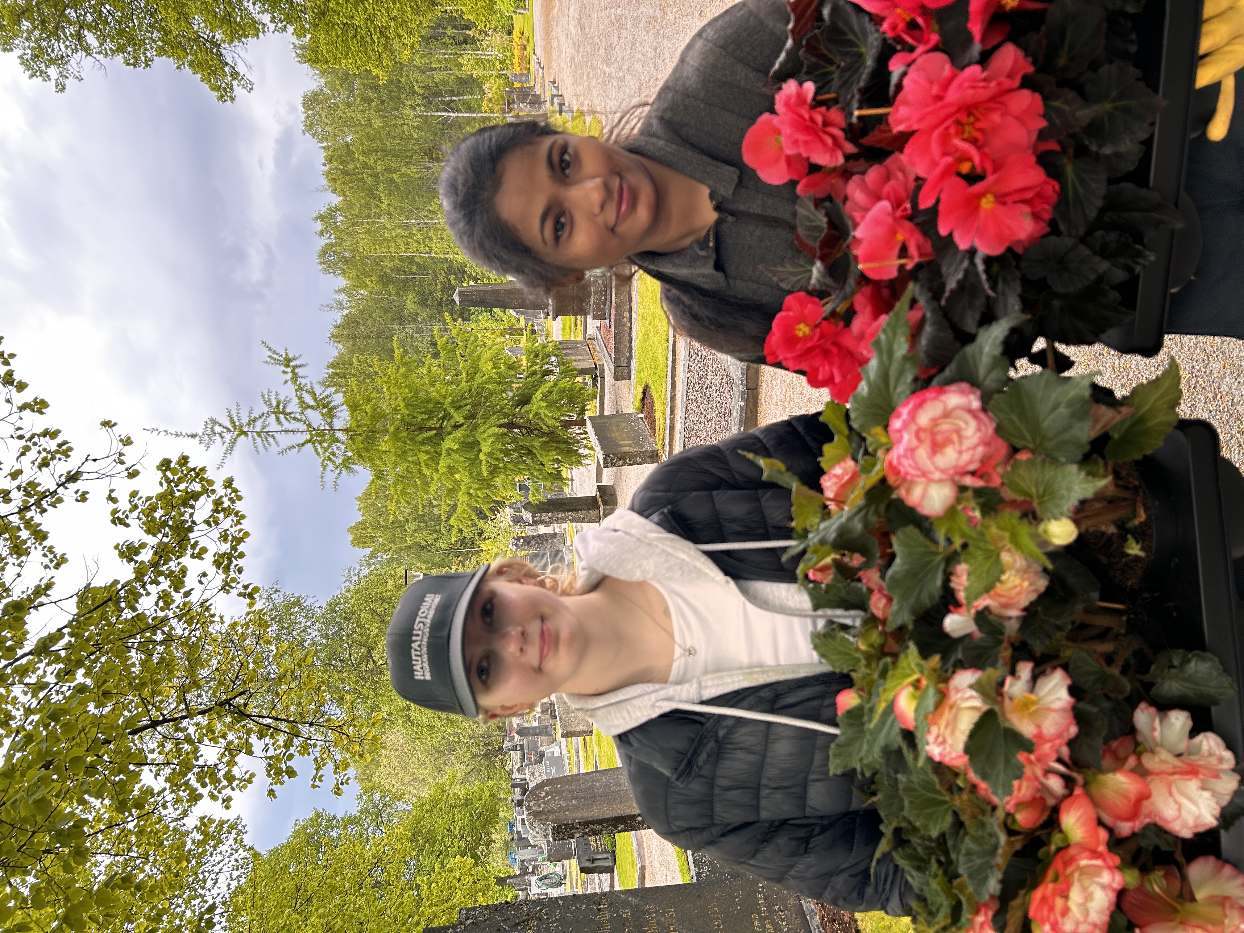 Två unga kvinnor på en begravningsplats håller lådor med blommor i sina famnar.