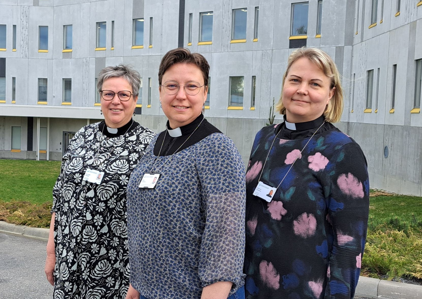 Tre kvinnor med prästkrage framför en grå cementbyggnad.